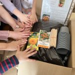 atvērta kartona kaste ar stādu trauciņiem, zemi un sēklu paciņām, virs tām bērnu rokas kas sniedzas kastē